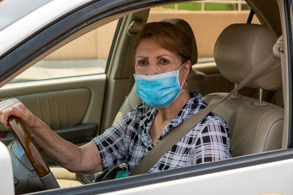 An older woman wearing a face mask sits in a car, gripping the steering wheel. 