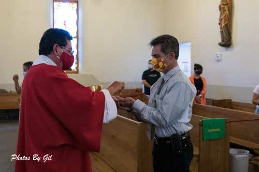 A priest in red robes gives communion to a man wearing a patterned mask in a church.
