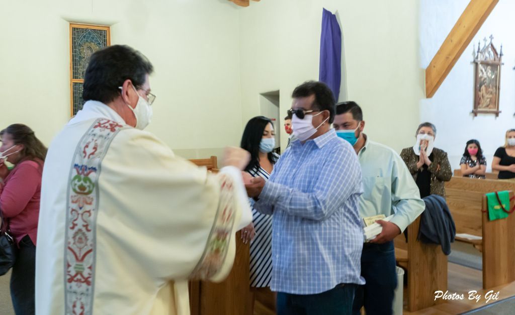 A priest in white robes, wearing a mask, hands a consecrated host to a masked man during a church service.