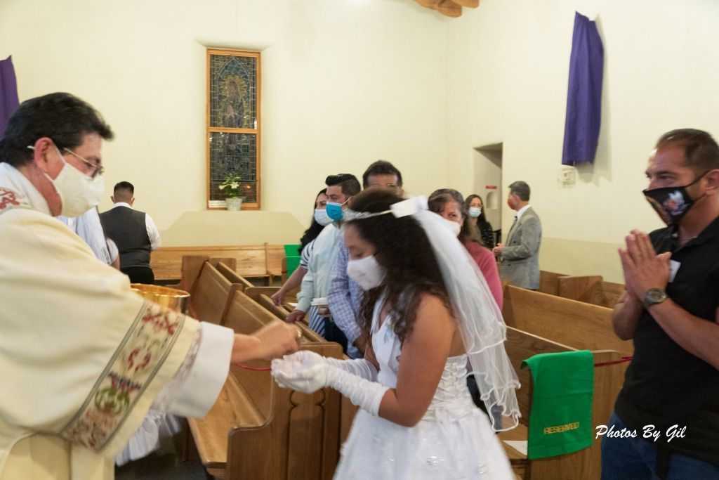 A priest in a decorated robe gives communion to a young girl in a white dress and veil. 