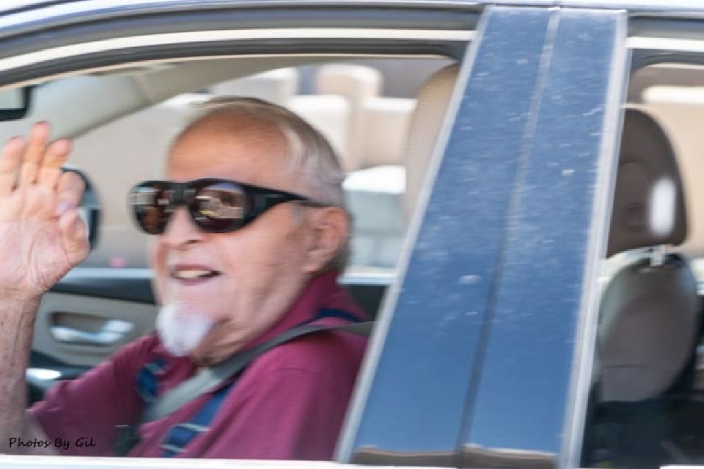 An elderly man with sunglasses and a goatee smiles warmly, waving from the passenger seat of a car. 