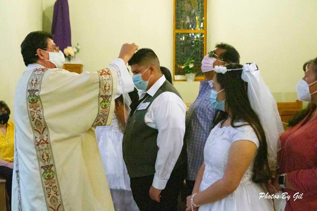 A priest in ornate vestments performs a blessing on two masked individuals in a church. 