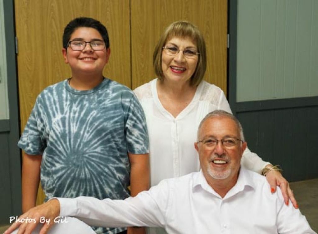 Smiling trio posing indoors, featuring a boy in a tie-dye shirt, a woman in a white blouse, and a seated man in a white shirt.