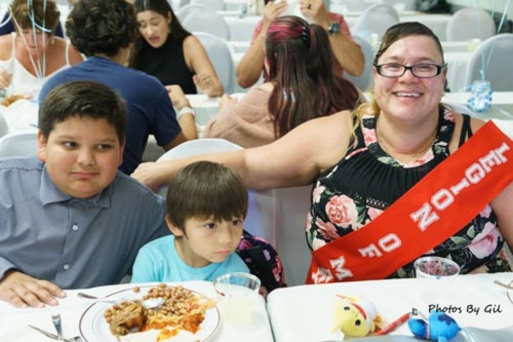 A smiling woman with a red sash sits at a table with two young boys.