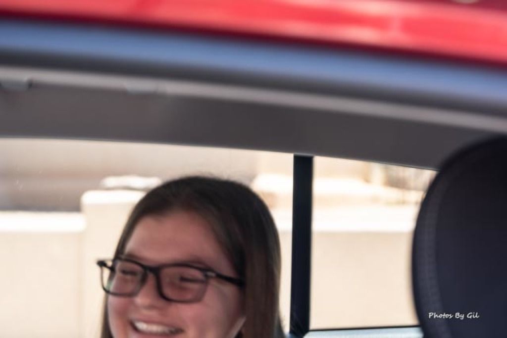 A close-up, slightly blurred image of a person wearing glasses, smiling inside a car.
