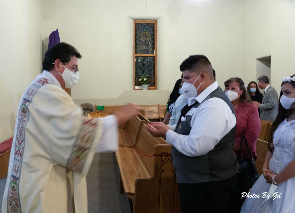 A priest in white robes gives communion to a man wearing a gray vest, both in masks.