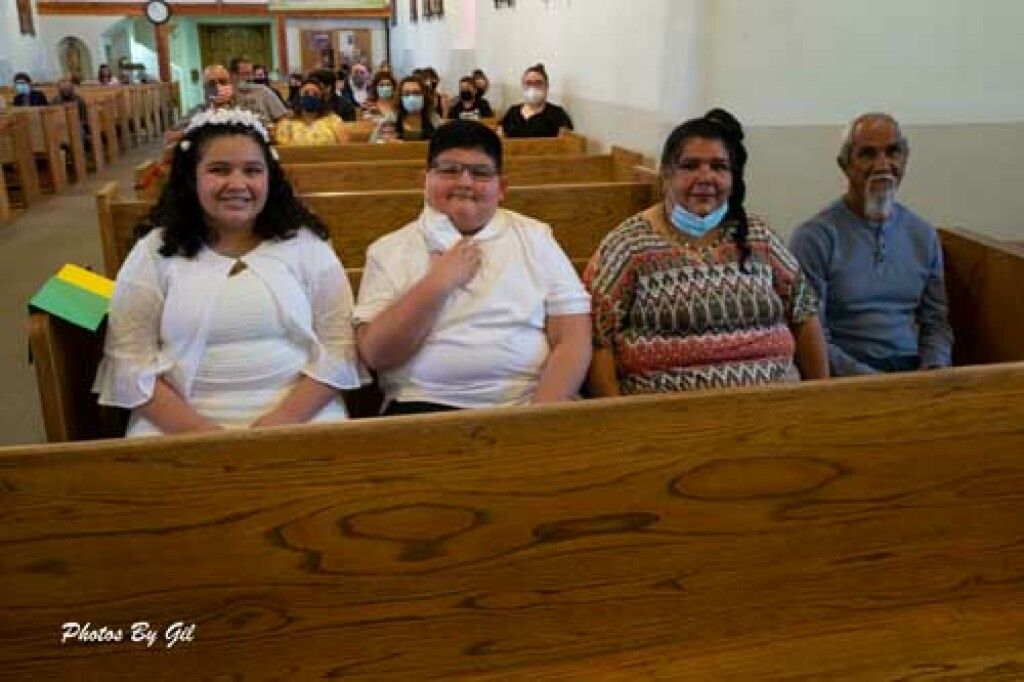 A young girl and boy, dressed in white, sit smiling in a church pew with two adults.