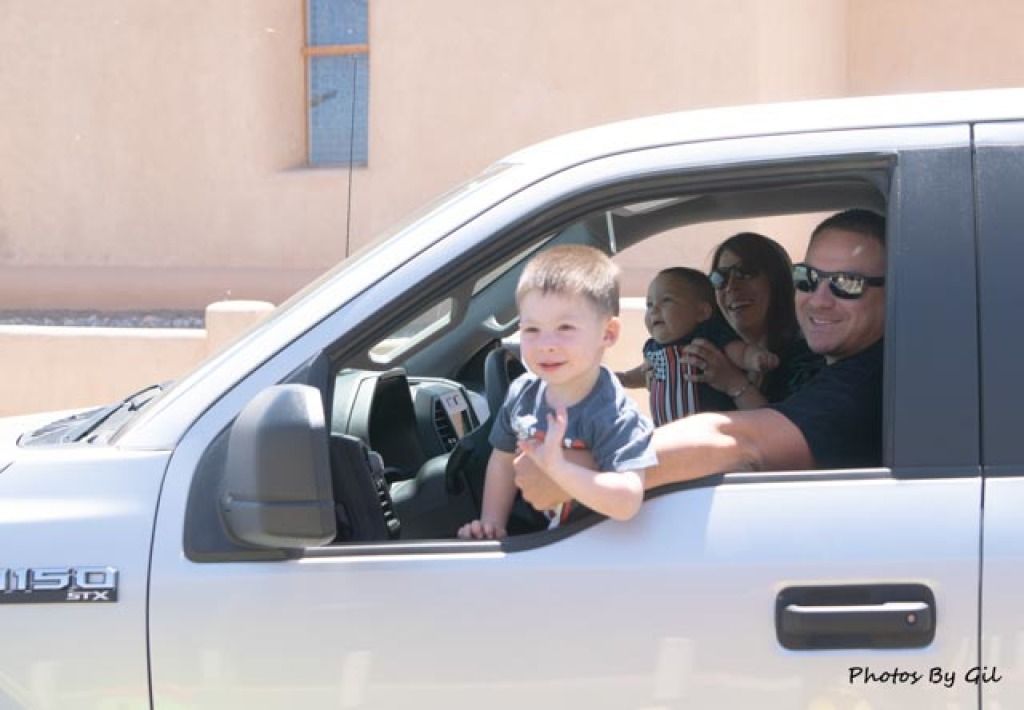 A young boy happily leans out of a white truck window on a sunny day.