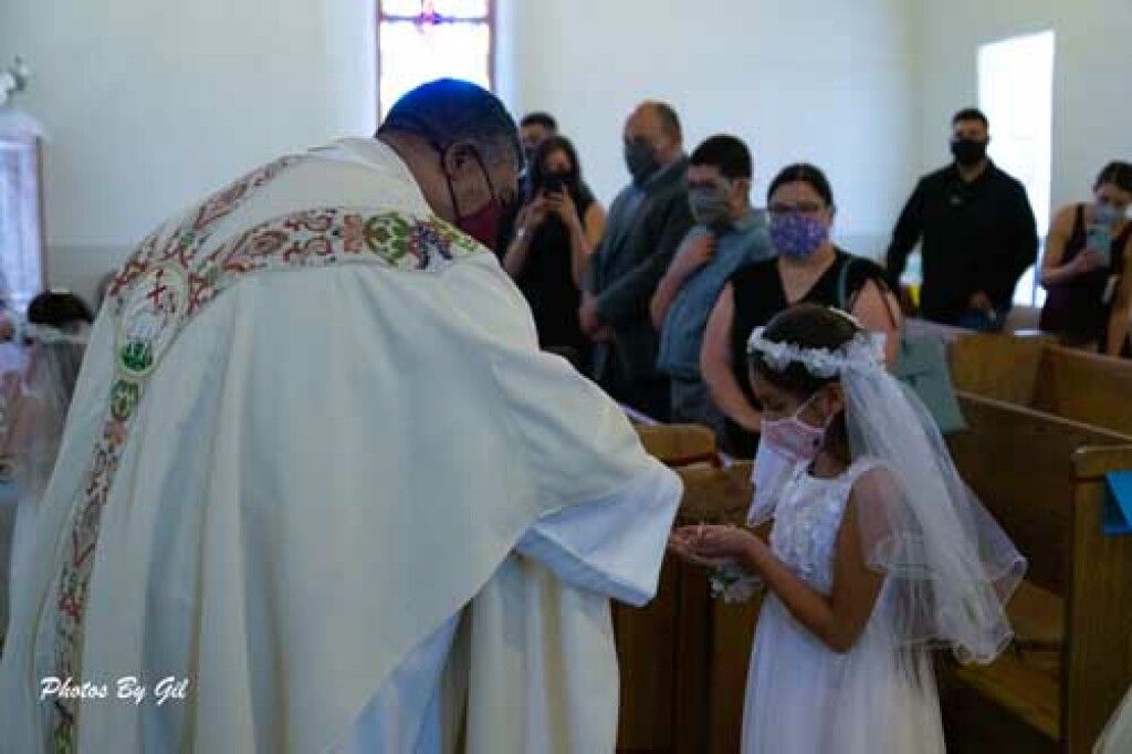 A priest in ornate robes gives communion to a young girl in a white dress and veil.