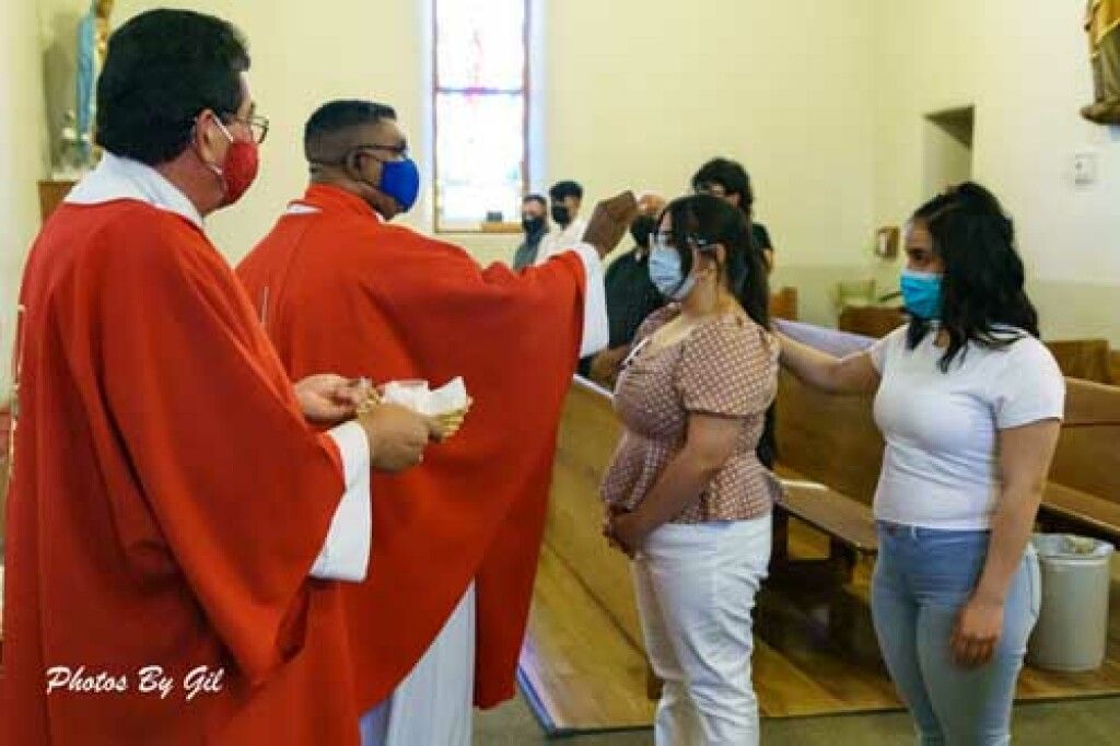 A priest wearing a red robe and mask performs a blessing on a masked woman in a church.