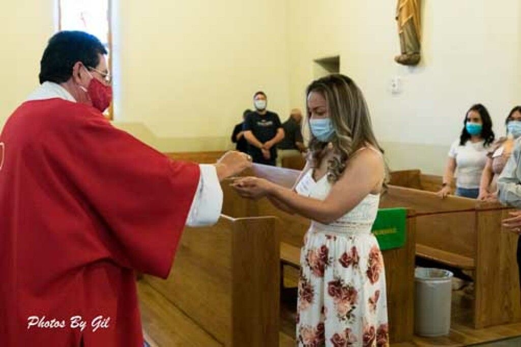 A priest in a red robe and face mask gives communion to a masked woman in a floral dress in a church.