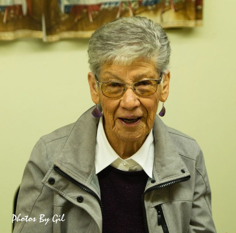 Elderly woman smiling warmly, wearing glasses and a gray jacket.