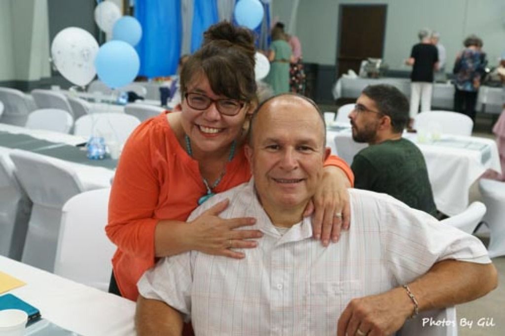 A smiling woman in an orange top stands behind a seated man in a white shirt at a festive event.