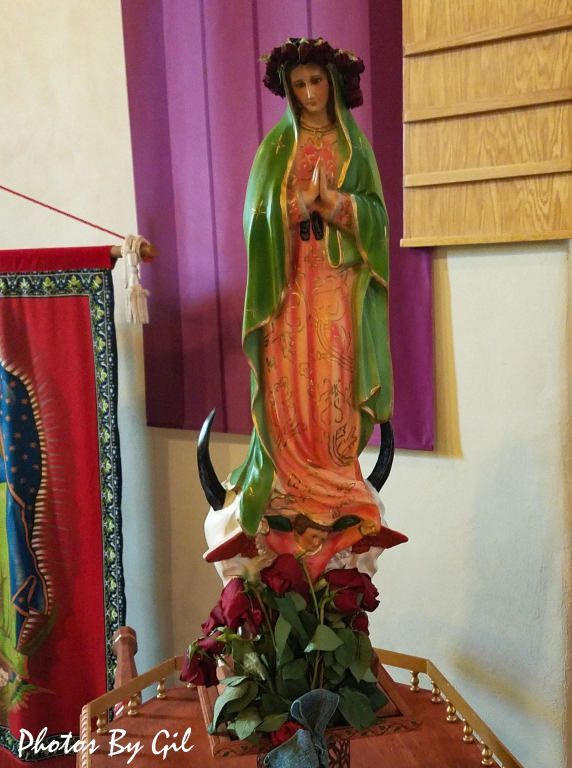 A statue of the Virgin Mary wearing a green mantle and pink dress, with a wreath of roses.