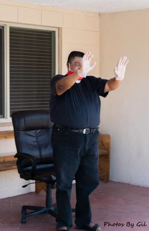 A person in a black outfit and white gloves stands on a porch, gesturing with both hands up. 