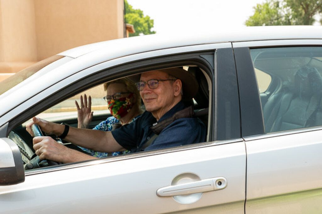 A smiling man wearing glasses and a hat sits in the driver's seat of a silver car. 