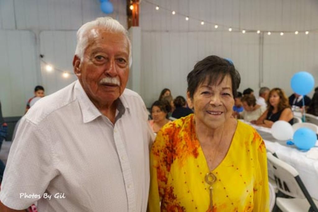 Elderly couple smiling at a festive indoor gathering, surrounded by people at tables with blue balloons and string lights.