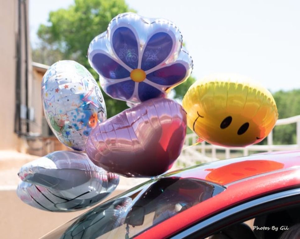 A cluster of colorful balloons, including a smiley face, starry pattern, and flower, float above a red car.