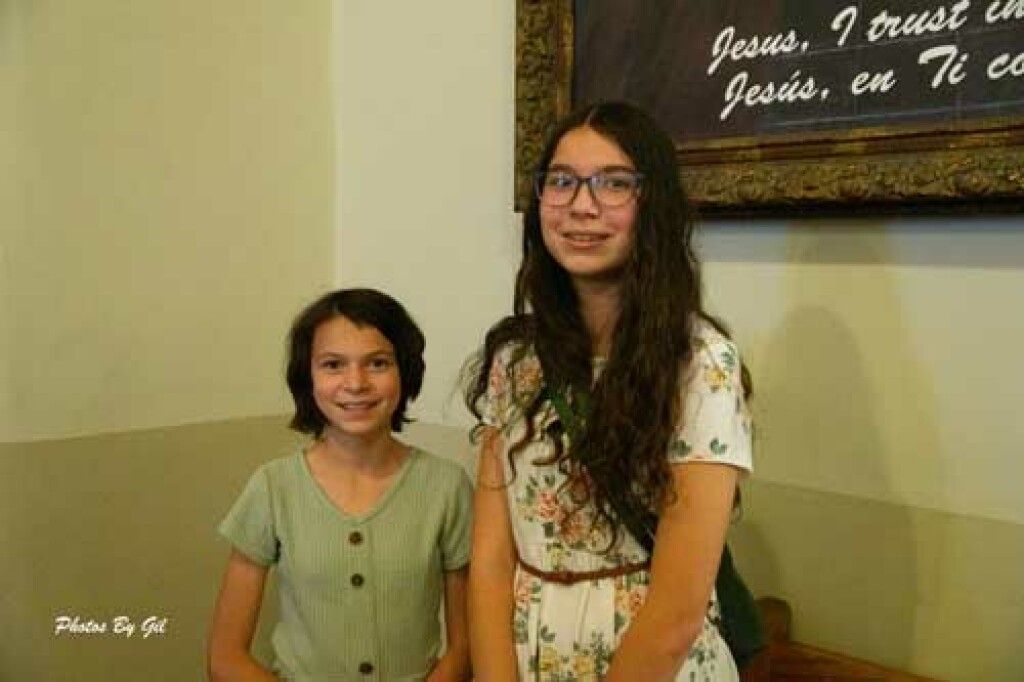 Two smiling girls stand indoors against a light wall, with one wearing a green dress and the other in a floral dress and glasses. 