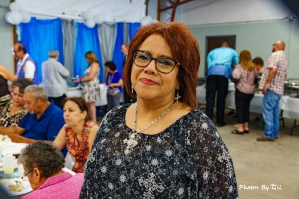 A woman in a patterned blouse and glasses smiles at the camera in a banquet hall. 