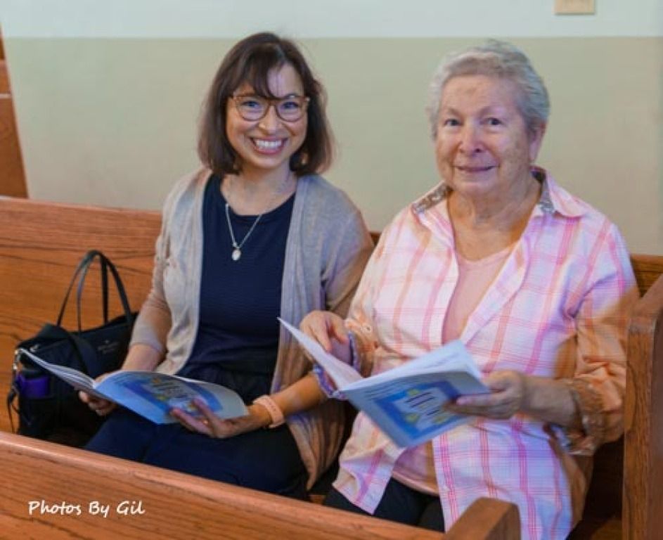 Two women sitting on a wooden pew, smiling, holding open hymnbooks. 