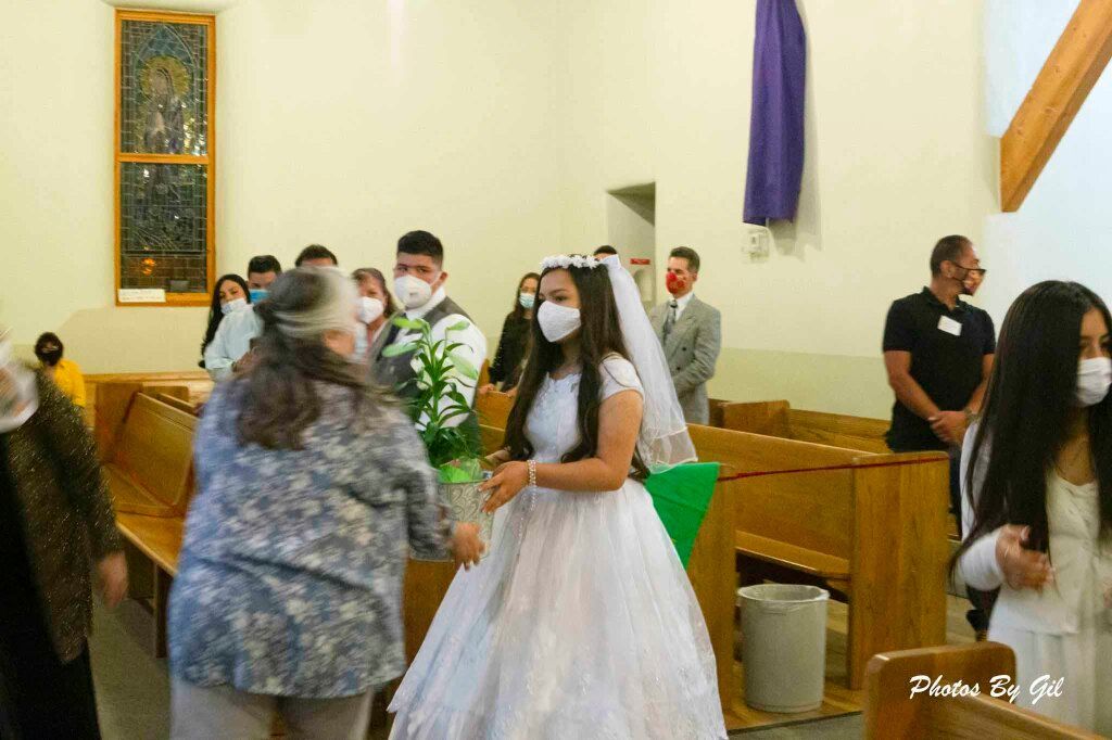 Young girl in a white dress and veil, holding a plant, walks down a church aisle.