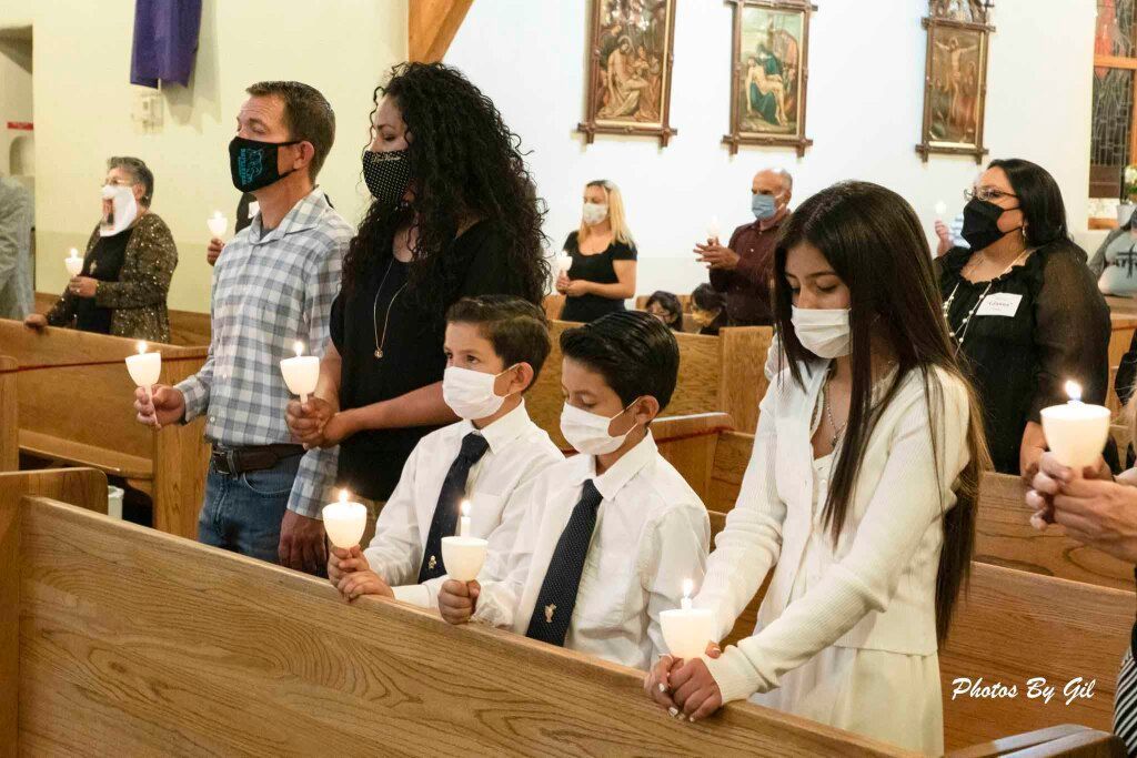People, including children, wearing masks and holding candles, stand solemnly in a church during a ceremony.
