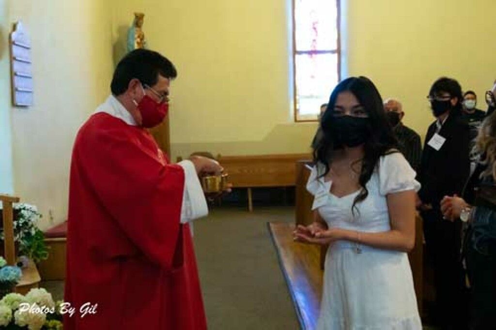 A priest in a red robe gives communion to a woman in a white dress at a church service. 