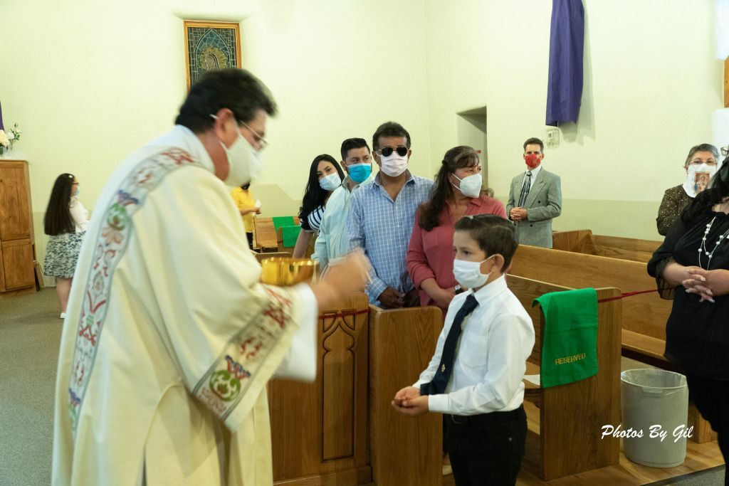 A priest wearing a mask in a church hands communion to a masked boy in a tie. 