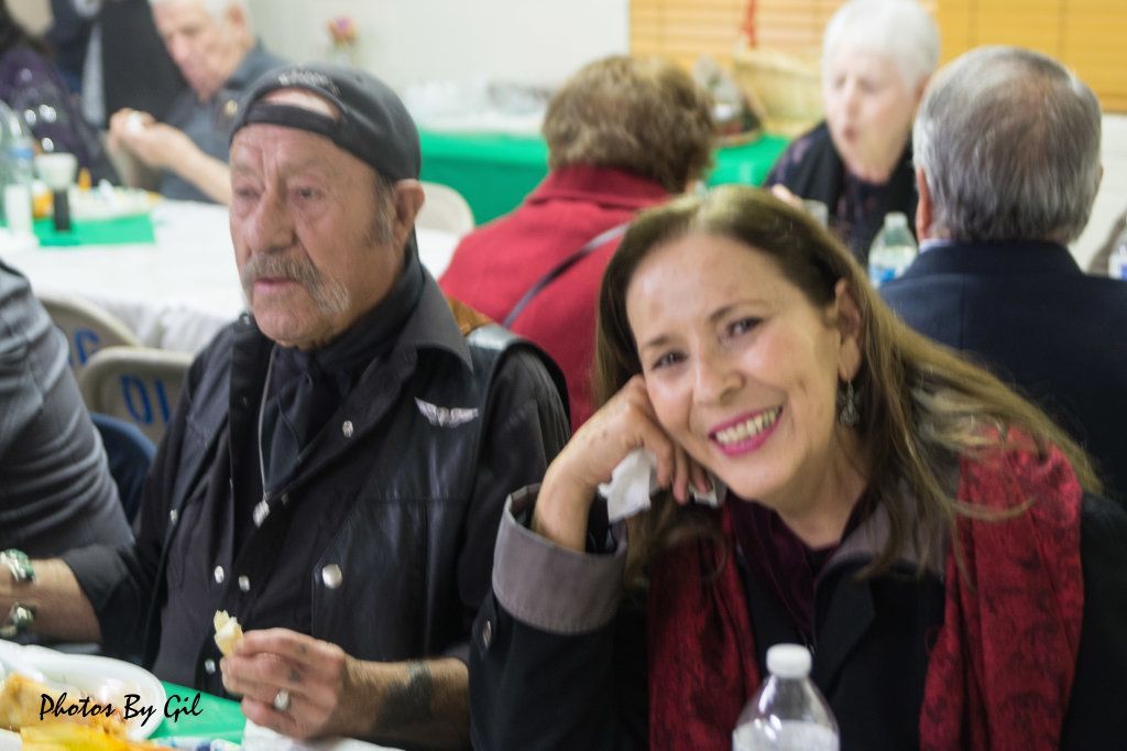 A woman in a red scarf smiles, leaning on one hand, while a man in a leather jacket and cap sits beside her.