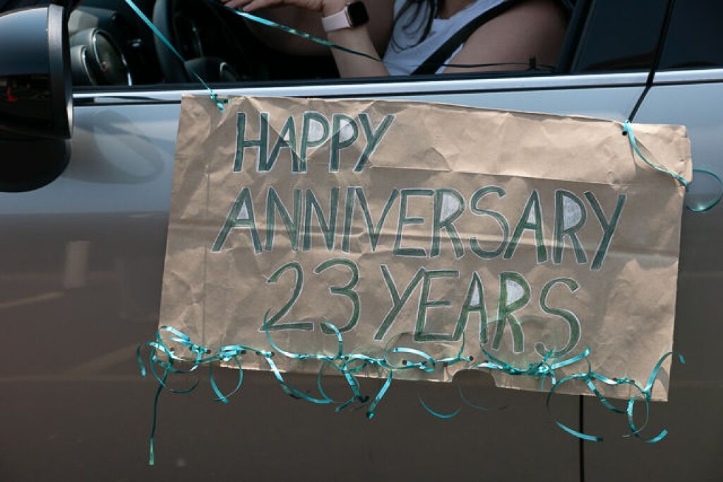 A masked person is in a car, holding a Happy Anniversary sign.
