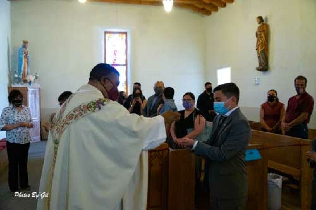 A priest in white robes, wearing a mask, performs a blessing for a masked man in a suit in a church.