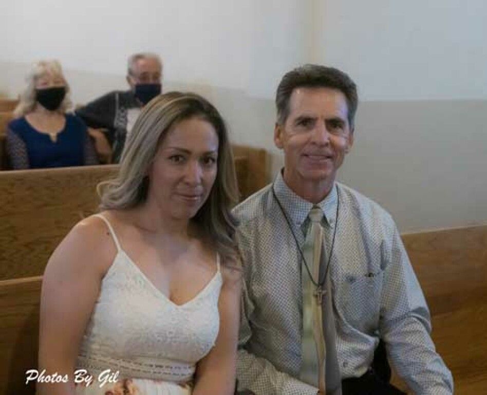 A woman in a white dress sits beside a man in a tie, smiling in a church pew.