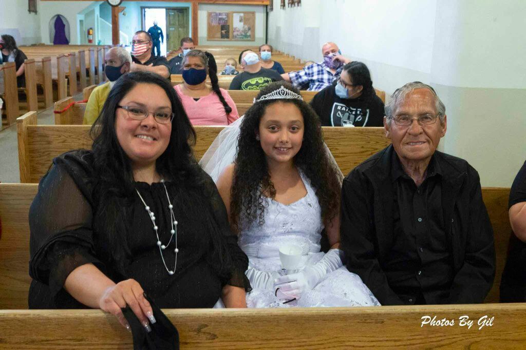 A smiling woman, girl in a white dress and tiara, and elderly man sit in a church pew.