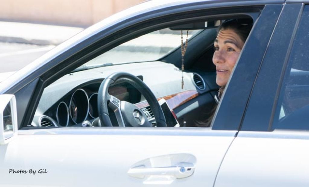 A woman is sitting in the driver's seat of a white car, smiling slightly, with a rosary hanging from the rearview mirror.