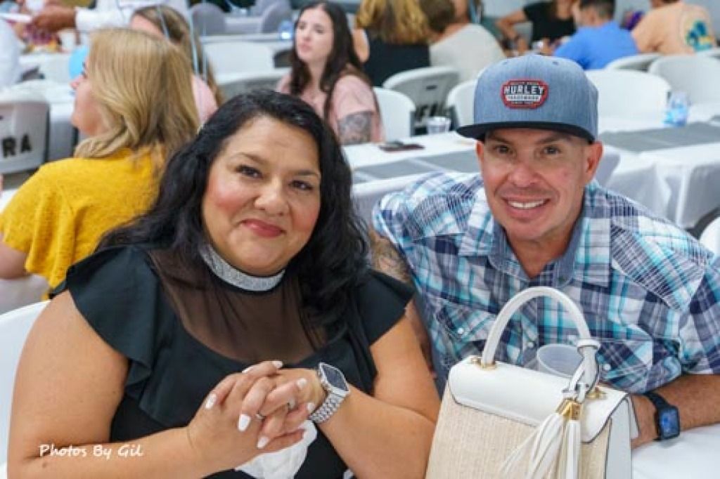 A smiling woman in a black dress and a man in a plaid shirt and cap sit at a table during a social gathering. 