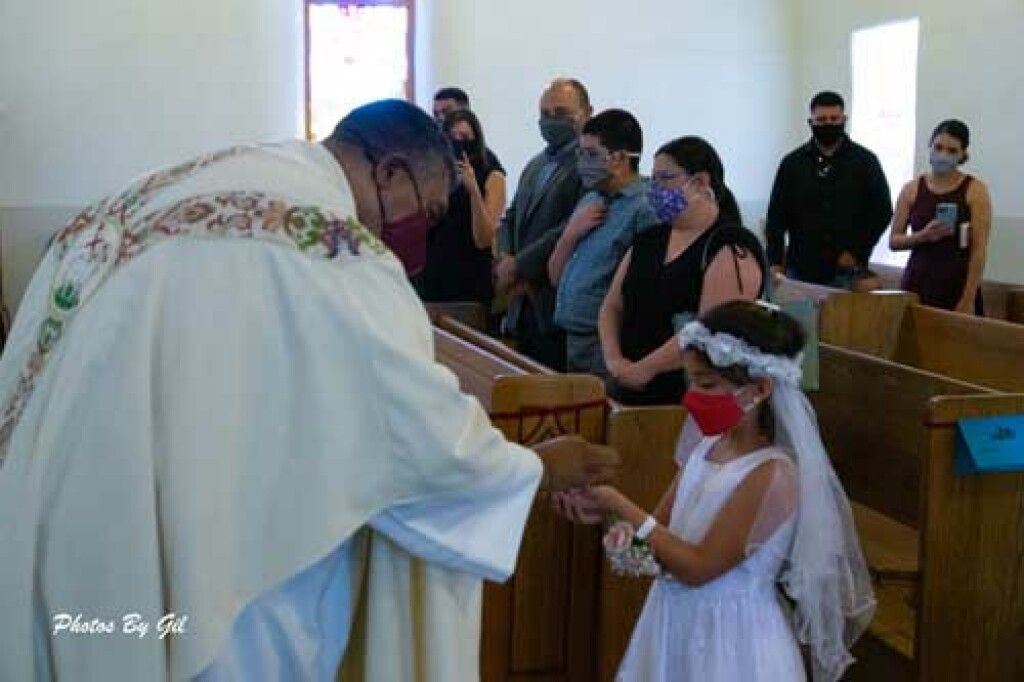 A priest hands communion to a young girl in a white dress and veil at a church.