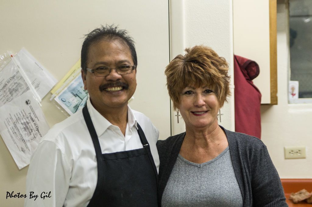A smiling man in an apron and glasses stands beside a woman with short hair and a gray sweater. 