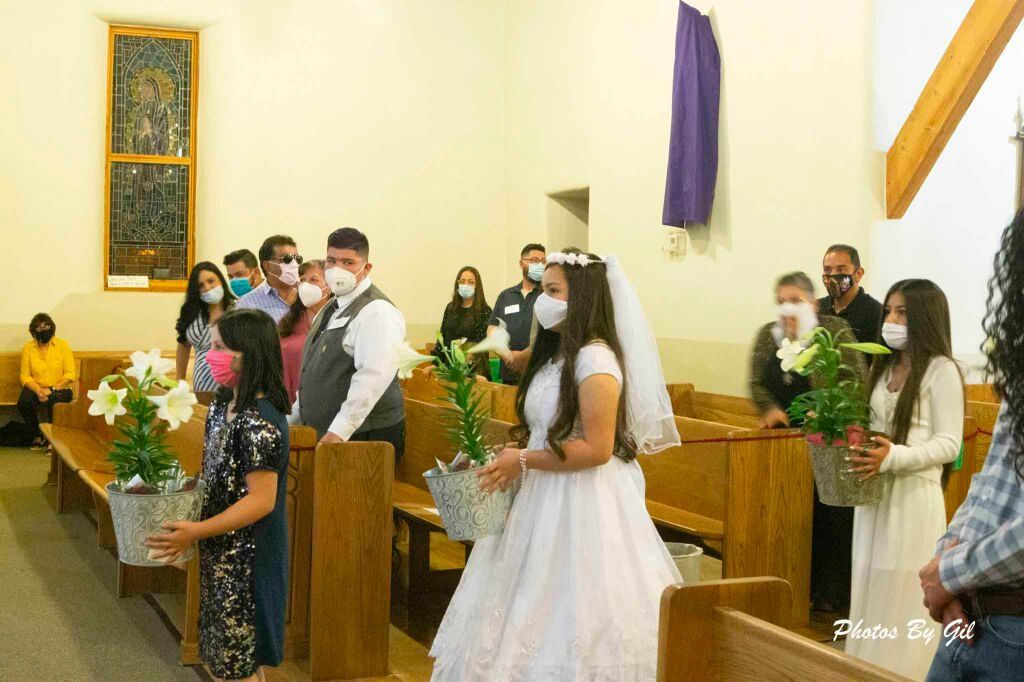 A young girl in a white dress and veil, wearing a mask, carries a potted plant in a church.