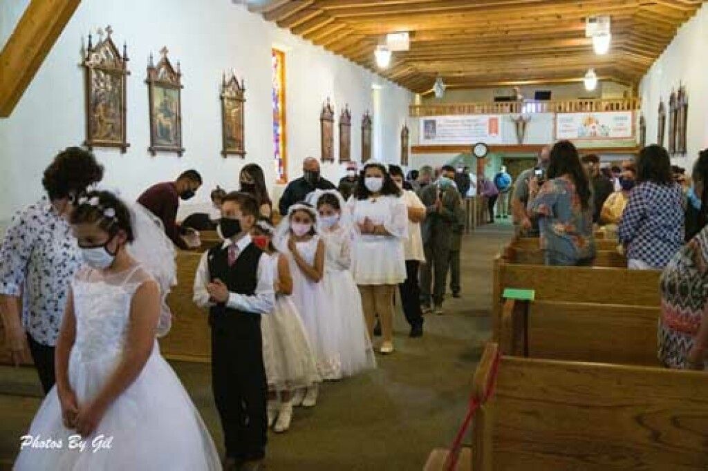 Children in formal attire, wearing masks, walk down a church aisle.