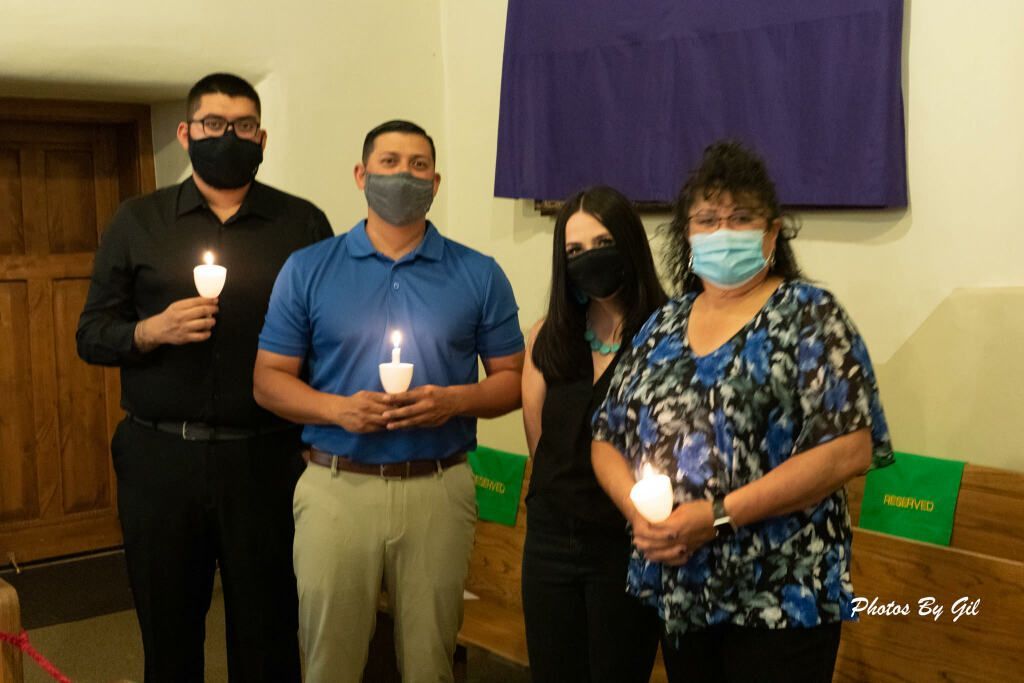 Four people wearing masks stand indoors, each holding a lit candle. 