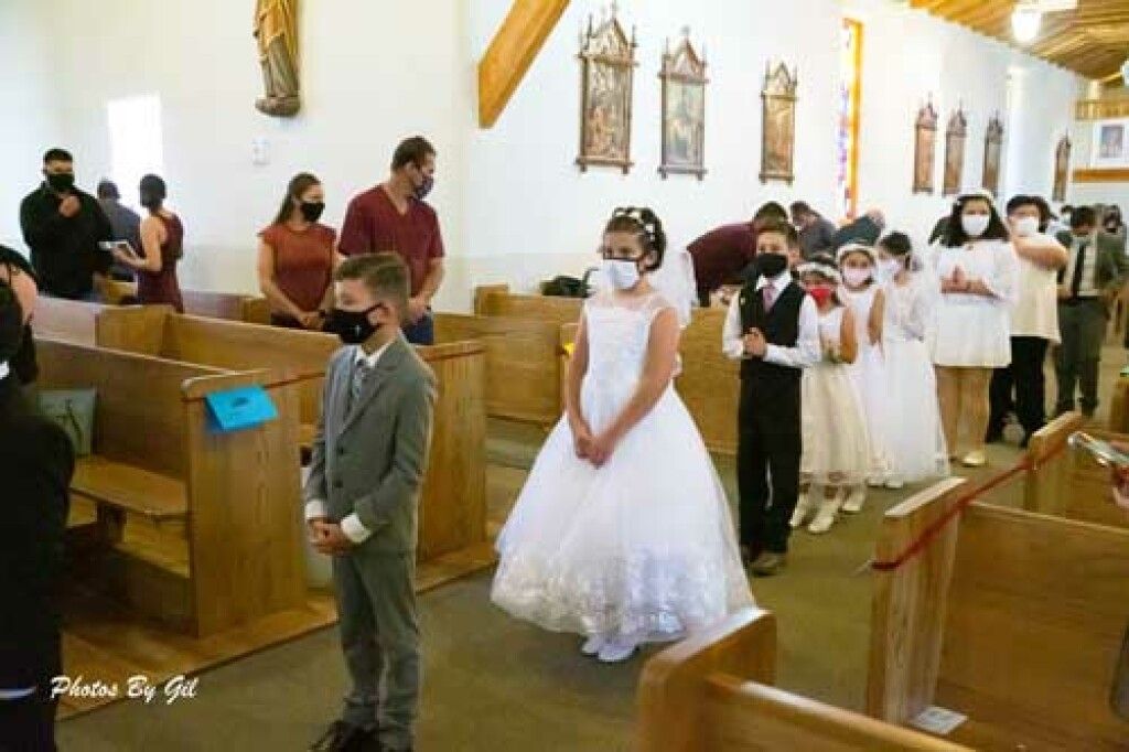 Children in formal attire, wearing masks, stand in a church aisle.