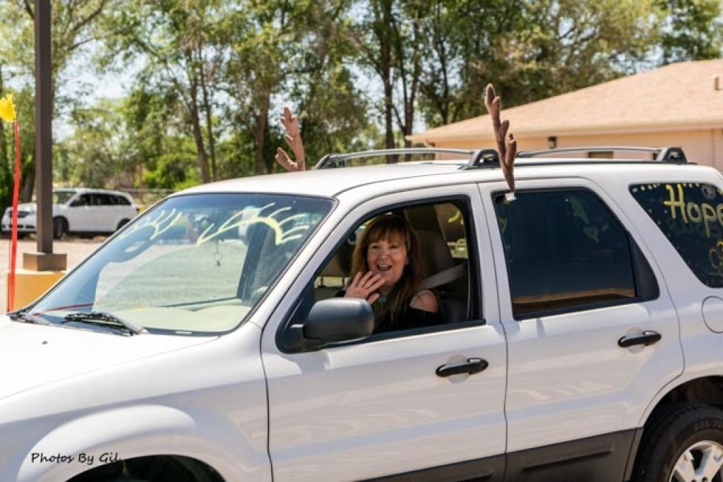 A woman waves happily from the driver's seat of a white SUV decorated with antlers and painted messages.
