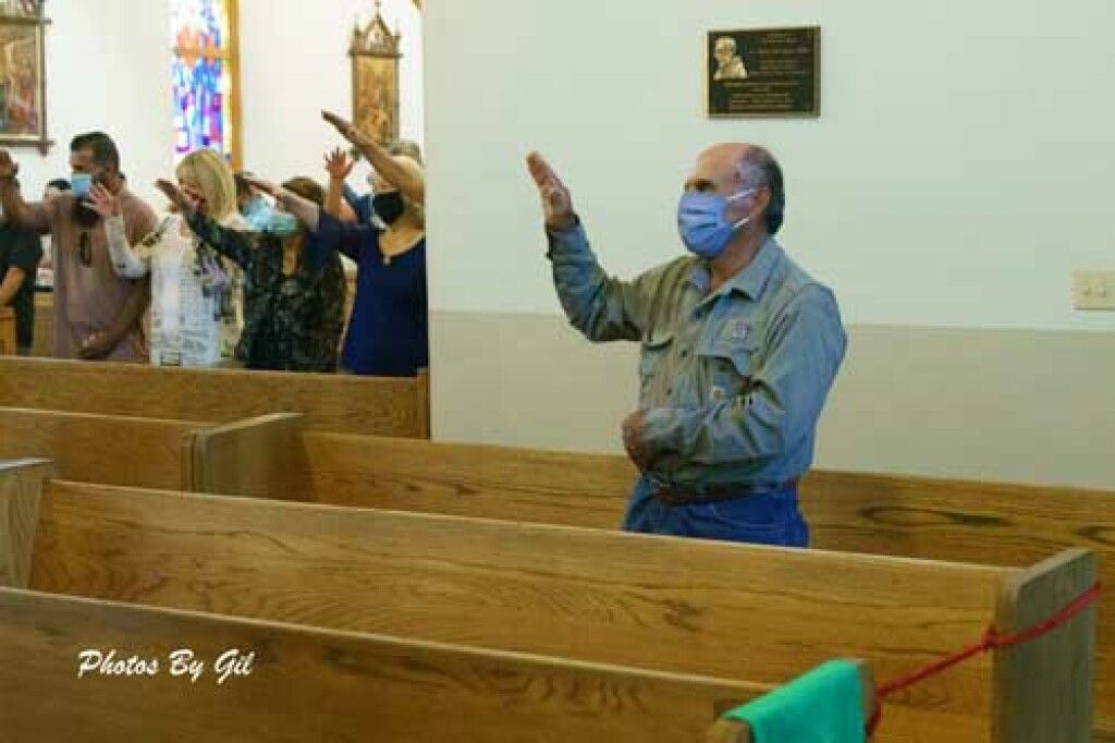 A diverse group of people, masked and socially distanced, stand in church pews raising their hands in a worship gesture. 