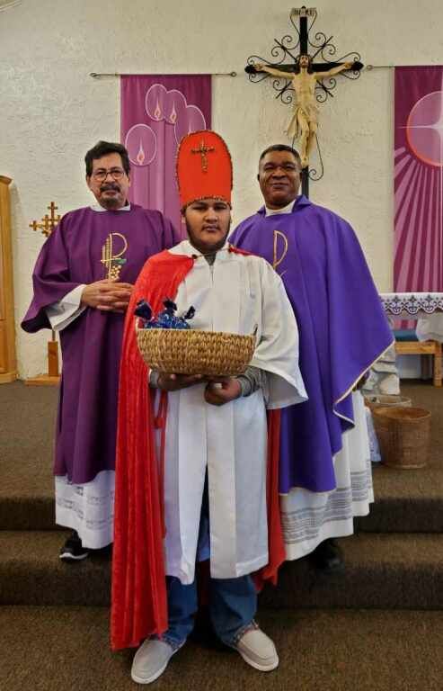 Three men stand in a church, two in purple robes and one in a white robe and red headpiece.