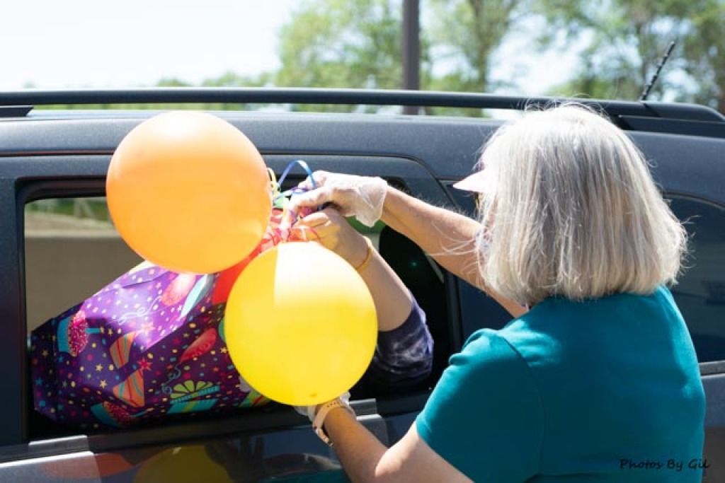 A woman with gray hair hands a large, colorful gift bag with orange and yellow balloons through a car window.