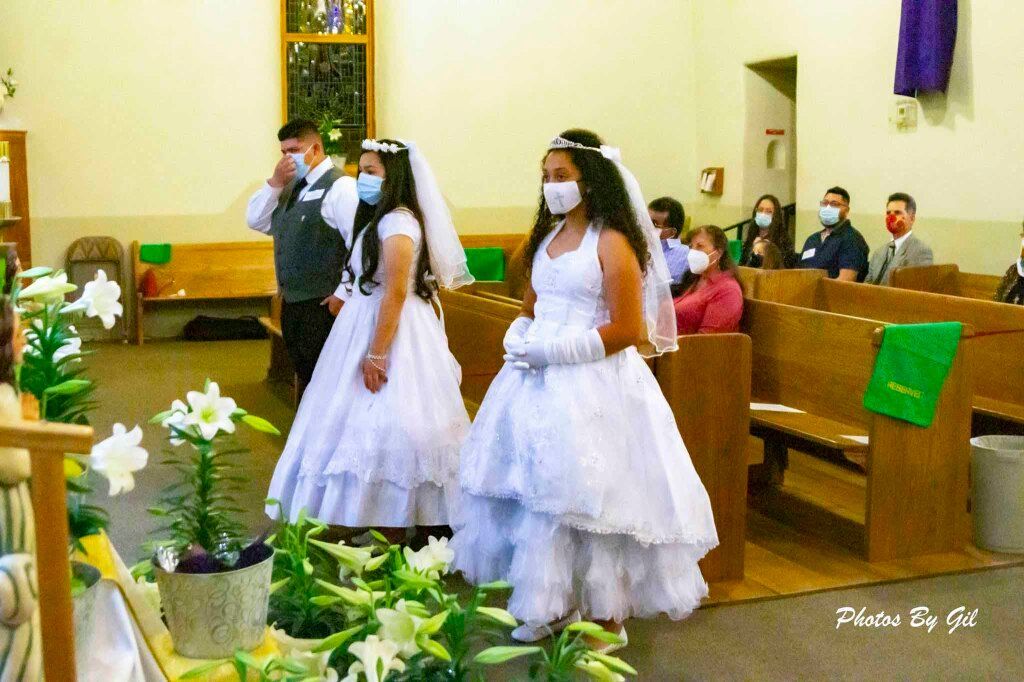 Two girls in white dresses and veils, wearing masks, stand in a church with a flower arrangement in front of them.