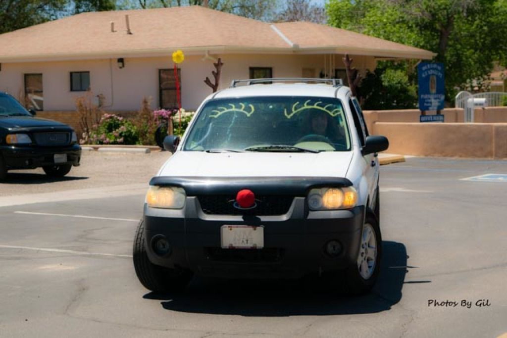 White SUV with a red nose and antlers, resembling a reindeer, parked in a sunny lot.