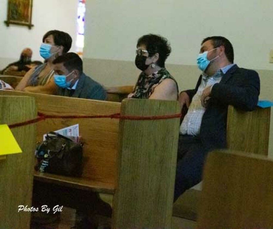Four people wearing face masks sit in wooden church pews, socially distanced, with solemn expressions.