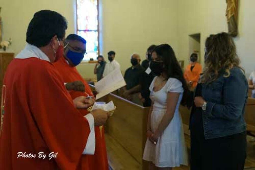 A priest in red robes reads from a paper in a church, facing a young woman in a white dress and a woman in a denim jacket.